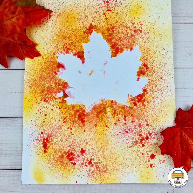 A piece of paper splattered with paint with a white space in the middle in the shape of a leaf. 